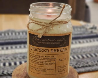 May include: A lit, glass jar candle with a brown label that reads "Charred Embers." The candle is tied with twine and sits on a small wooden stand. The label includes the text "Hand Poured Wood Wick Candles."