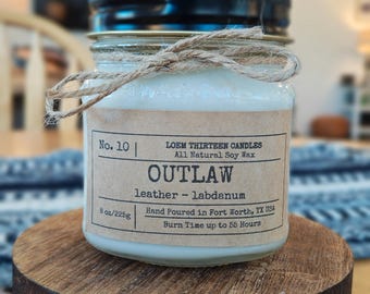 May include: A soy wax candle in a glass jar with a black lid, labeled "OUTLAW" with "leather - labdanum" scent. The label also indicates 8 oz/225g and hand-poured in Fort Worth, TX. The jar is tied with twine and sits on a wooden stand.