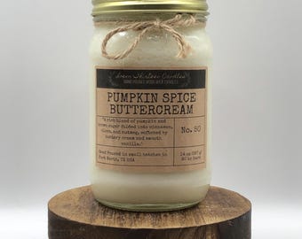 May include: A glass jar candle with a gold lid and a label that reads "Pumpkin Spice Buttercream." The candle is tied with twine and sits on a small, round wooden stand. The label includes the text "Loem Shirteen Candles."