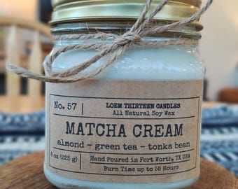 May include: A glass jar candle with a gold lid and a twine bow. The label reads "Matcha Cream" with scent notes and "8 oz (225 g)". The candle is hand-poured in Fort Worth, TX, and has a burn time up to 55 hours.