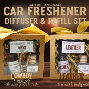 May include: Two car freshener diffuser and refill sets in cardboard boxes with gold ribbon. One set is labeled "Cowboy" with citrus, bergamot, and musk scent. The other is labeled "Leather" with resin, musk, and smoky wood scent.