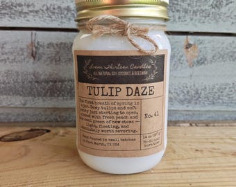 May include: A white candle in a glass jar with a gold lid and a twine bow. The label reads "Lrem Thirteen Candles," "TULIP DAZE," and "14 oz (397 g)." The text describes the scent as dewy tulips, soft peony, and fresh peach, noting a 75-80 hour burn time.
