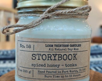 May include: A glass jar candle with a gold lid and a twine tie. The label reads "STORYBOOK" with "spiced honey - tonka" scent. The candle is 8 oz (225 g) and made with all-natural soy wax.