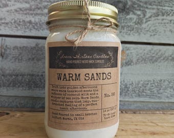 May include: A 14 oz glass jar candle with a gold metal lid and a twine bow. The label reads Loem Thirteen Candles, Warm Sands, No. 56, and includes text about teakwood, coconut milk, and sea salt, noting it is hand poured in Fort Worth, TX.
