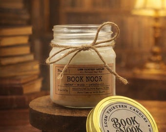 May include: A clear glass jar candle labeled "BOOK NOOK" with a brown label and a twine bow. The candle is on a small wooden stand, with a gold lid next to it. The candle is 6 oz (170g).