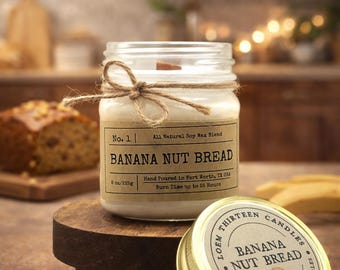 May include: A glass jar candle with a label that reads "BANANA NUT BREAD." The candle is tied with twine and sits on a wooden stand. The lid of a second candle is next to it. The label says "Hand Poured Wood Wick Candles."