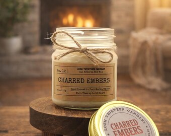 May include: A glass jar candle with a label that reads "CHARRED EMBERS" and a twine bow. The candle is on a small wooden stand, with the lid beside it. The lid has the words "CHARRED EMBERS" on it.