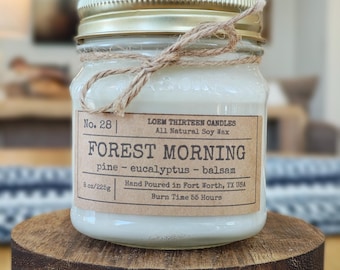 May include: A glass jar candle with a gold lid and a twine bow. The label reads "FOREST MORNING" with scent notes. The candle is 8 oz/225g and sits on a small, dark wooden stand. The text "LOEM THIRTEEN CANDLES" is also visible.