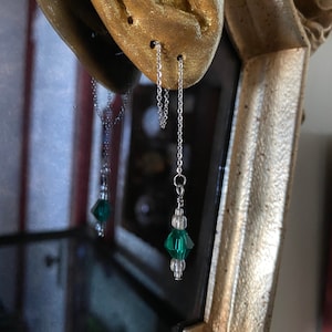 Pode incluir: Um par de brincos de corrente prateada com contas de cristal verde penduradas neles. Os brincos são mostrados pendurados em uma orelha modelo.
