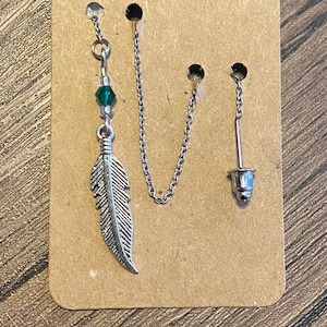 Pode incluir: Um brinco de prata com um pingente de pena, uma conta de cristal verde e uma corrente de prata. O brinco é apresentado em um cartão marrom.