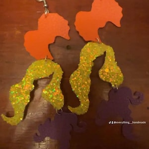May include: A pair of layered dangle earrings featuring three distinct shapes. The top piece is solid orange, the middle piece is yellow with a glitter finish, and the bottom piece is solid purple. Each earring hangs from a silver-toned metal hook.