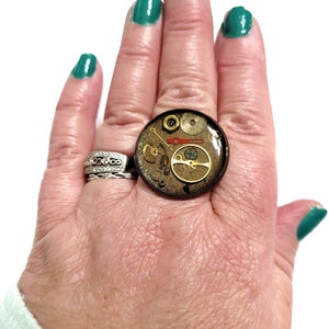 May include: A ring with a clear resin dome containing a variety of gold and silver gears and other watch parts. The ring is on a hand with green nail polish.