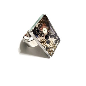 May include: A silver ring with a rectangular clear resin setting filled with assorted watch gears. The gears are in shades of gold and black.