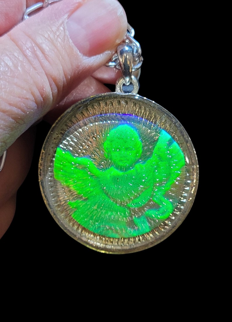 May include: A silver chain necklace with a round pendant featuring a green angel with iridescent wings. The pendant is encased in clear resin.