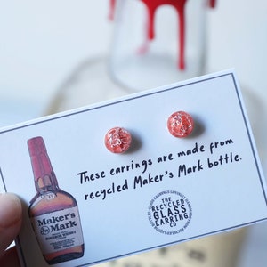May include: A pair of red and white stud earrings made from a recycled Maker's Mark bottle. The earrings are displayed on a white card with the text "These earrings are made from recycled Maker's Mark bottle." and a small image of a Maker's Mark bottle.