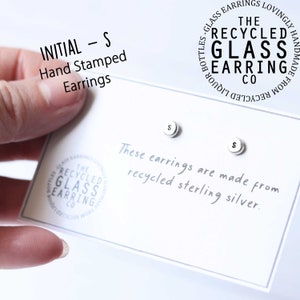 May include: A pair of sterling silver earrings with the letter "S" hand-stamped on each earring. The earrings are displayed on a white card with the text "These earrings are made from recycled sterling silver." and a logo for "The Recycled Glass Earring Co."