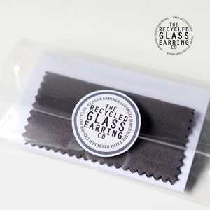 May include: A close-up of a small package containing a pair of gray fabric earring protectors. The package has a white label with the text "The Recycled Glass Earring Co. Lovingly Handmade from Recycled Liquor Bottles - Glass Earrings".