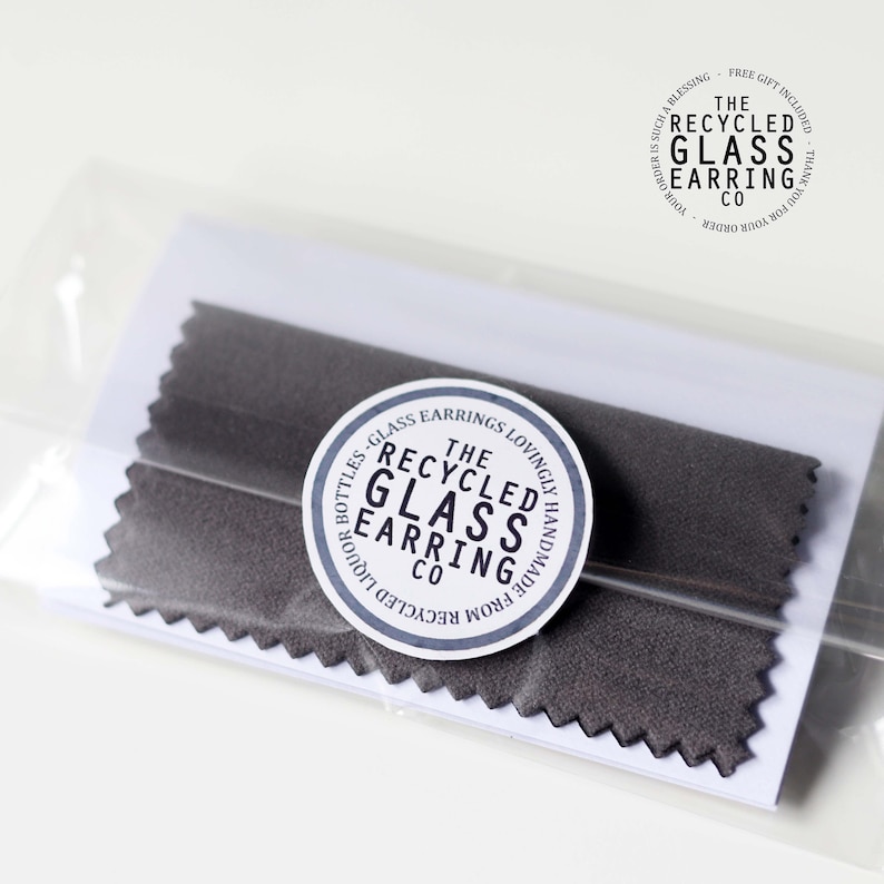 May include: A close-up of a small package containing a pair of gray fabric earring protectors. The package has a white label with the text "The Recycled Glass Earring Co. Lovingly Handmade from Recycled Liquor Bottles - Glass Earrings".