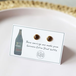 Peut inclure: Une paire de boucles d'oreilles en verre brun faites &agrave; partir d'une bouteille de Guinness Extra Stout recycl&eacute;e. Les boucles d'oreilles sont pr&eacute;sent&eacute;es sur une carte blanche avec un dessin d'une bouteille de Guinness et le texte "Ces boucles d'oreilles sont faites &agrave; partir d'une bouteille de Guinness Extra Stout."