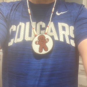 May include: A white and red beaded necklace with a round beaded pendant featuring a gingerbread man design. The necklace is worn by a person wearing a blue shirt with the word "COUGARS" printed on the front.