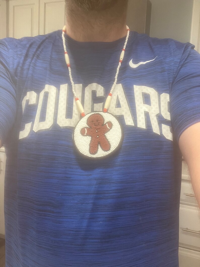 May include: A white and red beaded necklace with a round beaded pendant featuring a gingerbread man design. The necklace is worn by a person wearing a blue shirt with the word "COUGARS" printed on the front.