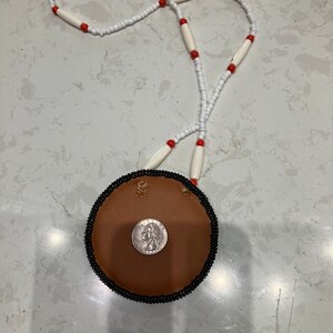 May include: A brown leather pouch with a black beaded trim and a silver coin inside. The pouch is attached to a white and red beaded necklace.