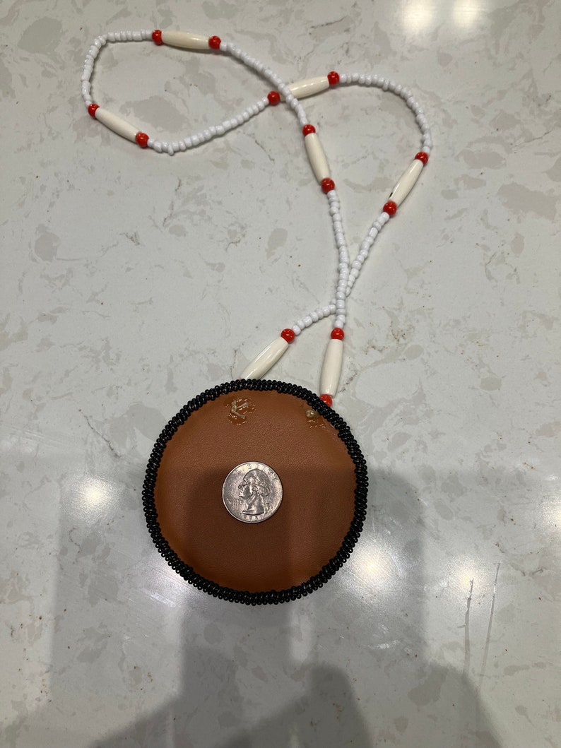 May include: A brown leather pouch with a black beaded trim and a silver coin inside. The pouch is attached to a white and red beaded necklace.