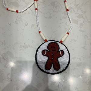 May include: A beaded necklace with a gingerbread man pendant. The pendant is made of white beads with brown and red beads forming the gingerbread man. The necklace is made of white beads with red beads interspersed.