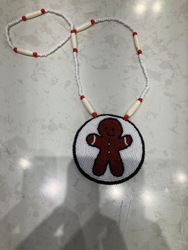 May include: A beaded necklace with a gingerbread man pendant. The pendant is made of white beads with brown and red beads forming the gingerbread man. The necklace is made of white beads with red beads interspersed.