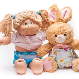 May include: A Cabbage Patch Kid doll with blonde hair and a pink shirt is sitting next to a brown plush bunny wearing a blue and white diaper. Both dolls are wearing blue and white patterned diapers.