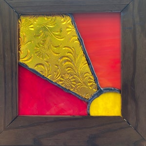 May include: A framed stained glass piece with a geometric design. The glass is in shades of red, yellow, and gold. The frame is dark brown wood.