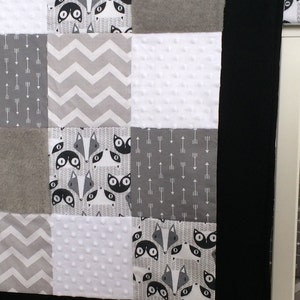 May include: A patchwork baby blanket with a black border. The quilt features squares of gray, white, and black fabrics. Some squares have fox faces, arrows, or chevron patterns. Other squares are solid gray or white with a textured dot pattern.