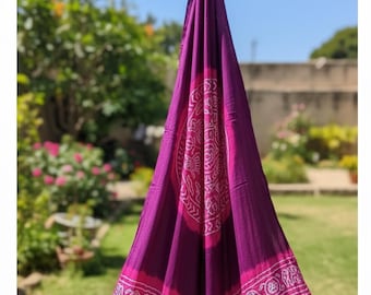 May include: A draped, vibrant purple and pink textile with intricate white patterns. The fabric features a central circular design and a border with elephant motifs. The textile is hanging outdoors, with a backdrop of greenery and a clear blue sky.