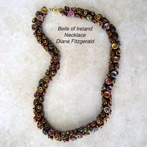 Pode incluir: Um colar de contas marrons, douradas e verdes com um fecho. O colar é feito de pequenas contas redondas que são dispostas em um padrão. O colar é chamado de "Bells of Ireland Necklace" e é de "Diane Fitzgerald".