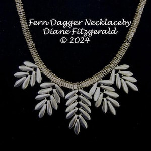 May include: A silver necklace with a fern-like design. The necklace is made of small, silver-toned beads that are arranged in a cascading pattern. The necklace is called "Fern Dagger Necklace" and is by Diane Fitzgerald. The copyright symbol and the year 2024 are also included in the image.
