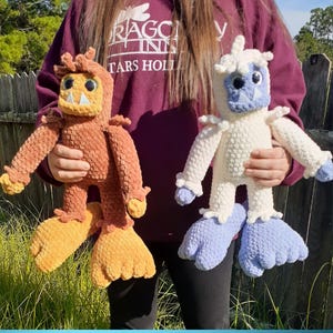 May include: Two handmade crochet monster dolls. One is brown and orange, the other is white and blue. Both have large feet, arms, and a face with a toothy grin. The dolls are held in front of a green background.