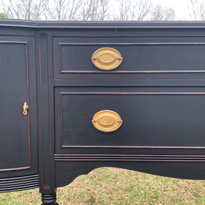 May include: A black painted antique sideboard with two drawers and a cabinet door. The drawers have oval brass pulls. The sideboard has a curved bottom and sits on four legs.