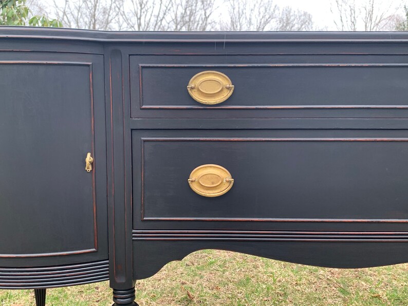 May include: A black painted antique sideboard with two drawers and a cabinet door. The drawers have oval brass pulls. The sideboard has a curved bottom and sits on four legs.