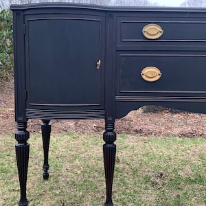 May include: A black painted wooden buffet with a curved top, two drawers, and a single door. The buffet has four turned legs and brass hardware.