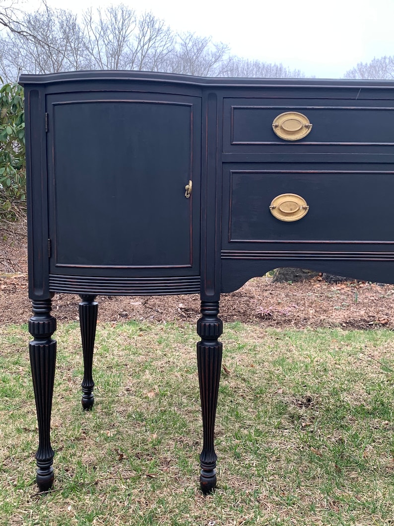 May include: A black painted wooden buffet with a curved top, two drawers, and a single door. The buffet has four turned legs and brass hardware.