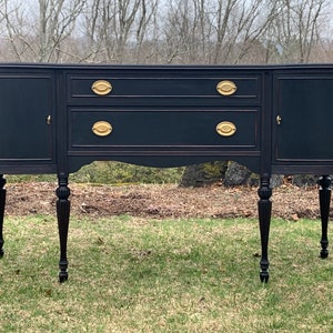 May include: A black painted wooden sideboard with gold hardware and turned legs. The sideboard has two doors on each end and two drawers in the center.