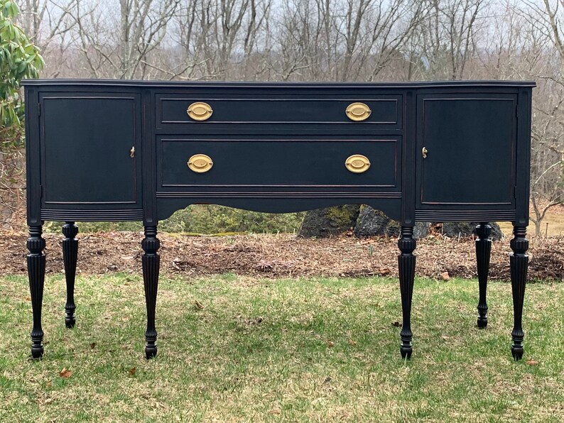 May include: A black painted wooden sideboard with gold hardware and turned legs. The sideboard has two doors on each end and two drawers in the center.
