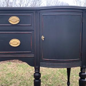 May include: A black painted antique sideboard with two drawers and a cabinet door. The cabinet door has a brass knob and the drawers have brass oval pulls. The sideboard has turned legs and a curved front.