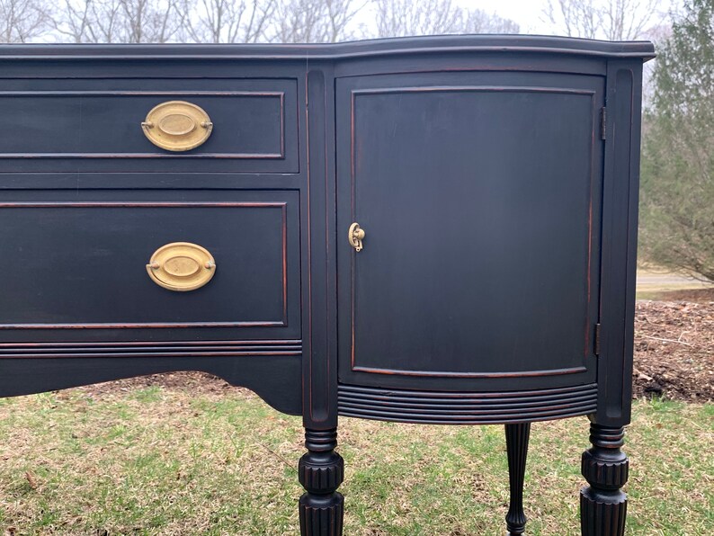 May include: A black painted antique sideboard with two drawers and a cabinet door. The cabinet door has a brass knob and the drawers have brass oval pulls. The sideboard has turned legs and a curved front.