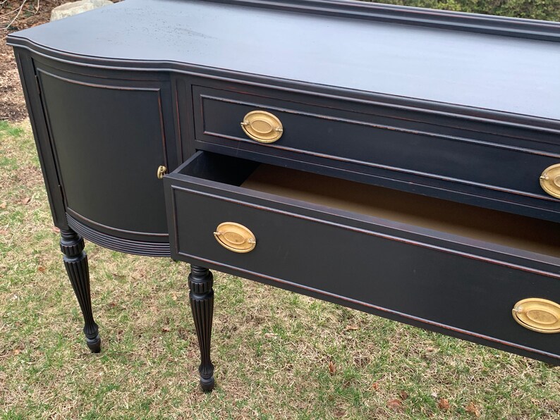 May include: Black painted wooden sideboard with three drawers and two cabinet doors. The sideboard has turned legs and brass hardware.