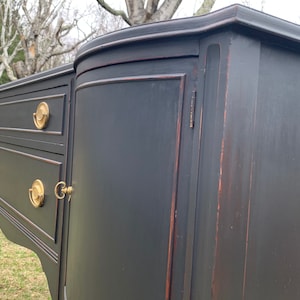 May include: A black painted antique sideboard with two drawers and a cabinet door. The cabinet door has a brass handle and keyhole. The sideboard has turned legs and a curved front.