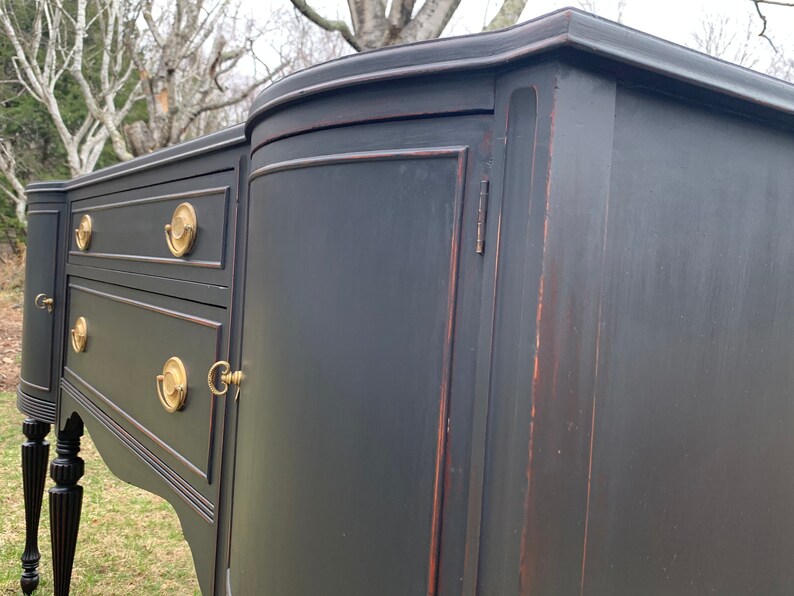May include: A black painted antique sideboard with two drawers and a cabinet door. The cabinet door has a brass handle and keyhole. The sideboard has turned legs and a curved front.