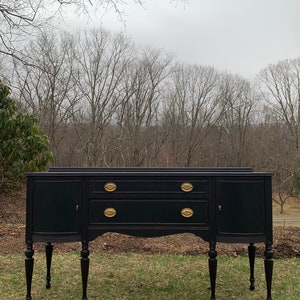 May include: A black painted vintage sideboard with gold hardware and turned legs. The sideboard has two doors and two drawers. It is sitting on a green lawn in front of a forest.