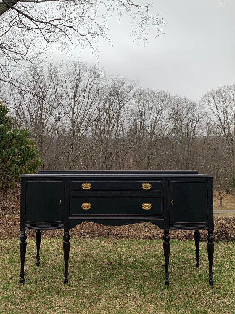 May include: A black painted vintage sideboard with gold hardware and turned legs. The sideboard has two doors and two drawers. It is sitting on a green lawn in front of a forest.