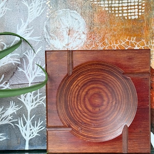 May include: A square, wooden art piece with a circular, carved design in the center. The wood has a rich, reddish-brown color. The background features abstract art with white and orange tones, and a hint of green foliage.